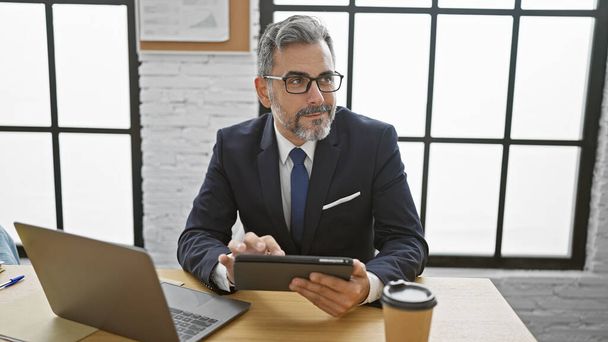 Homem de negócios hispânico jovem confiante com cabelos grisalhos, trabalhando em laptop e touchpad no escritório, sorrindo largo - Foto, Imagem
