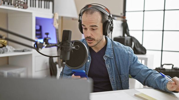 Hombre hispano con barba graba un podcast en un estudio moderno, usando un micrófono y un smartphone. - Foto, Imagen