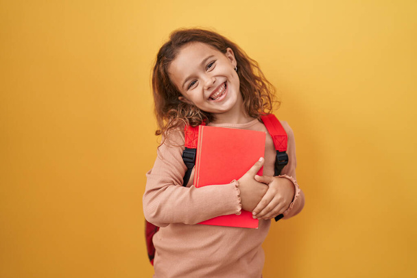 Pequeña chica caucásica con mochila de estudiante y celebración de libro logro con sonrisa feliz y la expresión del ganador con la mano levantada  - Foto, imagen