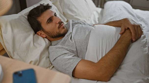 Удумливий іспаномовний чоловік лежить в ліжку, дивлячись в сторону в розслабленому приміщенні спальні, що представляє відпочинок і інтроспекцію. - Фото, зображення