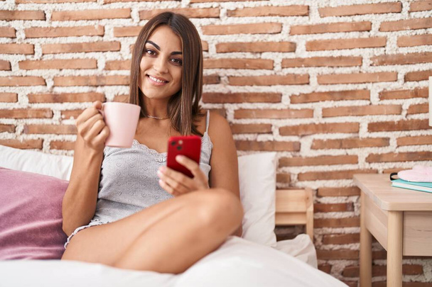 Genç ve güzel İspanyol kadın akıllı telefon kullanıyor. Yatak odasında kahve içiyor. - Fotoğraf, Görsel