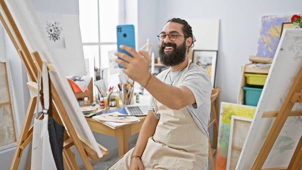 Homem hispânico com barba tomando selfie no estúdio de arte cercado por pinturas e cavaletes, exalando criatividade e alegria. - Foto, Imagem