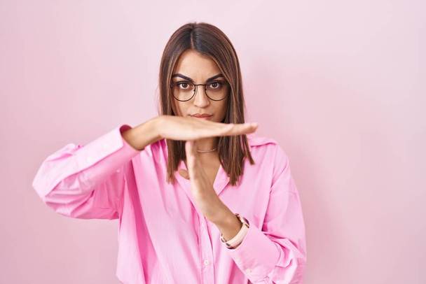 Νεαρή ισπανίδα γυναίκα φορώντας γυαλιά στέκεται πάνω από ροζ φόντο κάνει διάλειμμα από τα χέρια με τα χέρια, απογοητευμένος και σοβαρή πρόσωπο  - Φωτογραφία, εικόνα