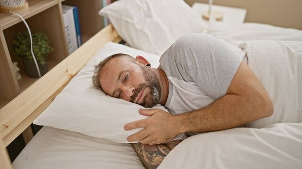 Красивый молодой человек лежит на уютной кровати, показывая серьезное выражение лица, отражая сосредоточенный образ жизни в хорошо освещенной спальне - Фото, изображение