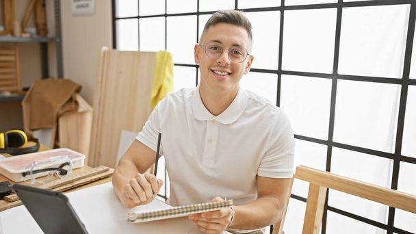 Ένας νεαρός Ισπανός με γυαλιά χαμογελά σε ένα καλά φωτισμένο εργαστήριο ξυλουργικής κρατώντας ένα σημειωματάριο.. - Φωτογραφία, εικόνα