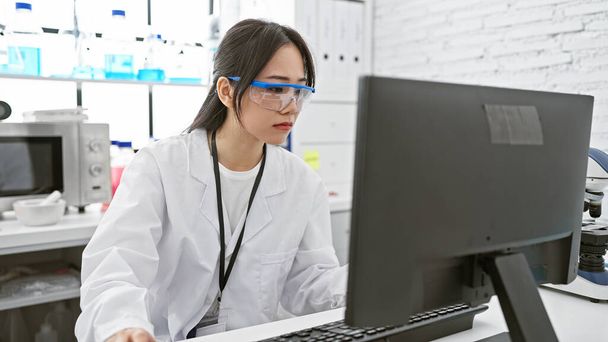 Uma jovem mulher asiática focada em um casaco de laboratório e óculos de segurança trabalhando em um computador em um laboratório moderno. - Foto, Imagem