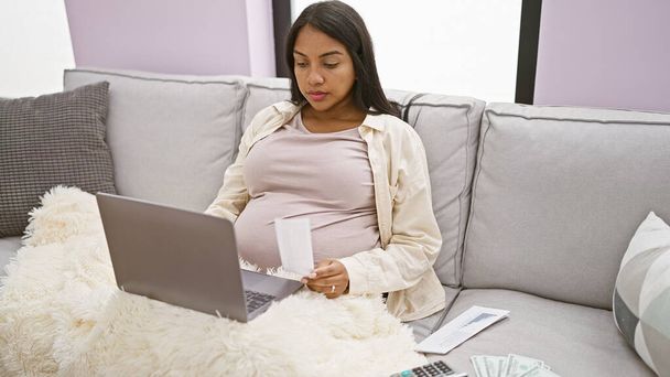 Oczekując młoda Latynoska kobieta wygodnie pracuje na laptopie księgowania gospodarki domowej z jej salonu sofa - Zdjęcie, obraz