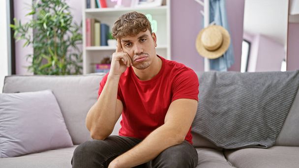 Portret van een serieuze jonge Spaanse man, zittend op een bank, denkend aan een probleem binnen. een knappe man die zich zorgen maakt in zijn woonkamer, weerspiegelt zijn droevige, twijfelachtige gedachten thuis. - Foto, afbeelding