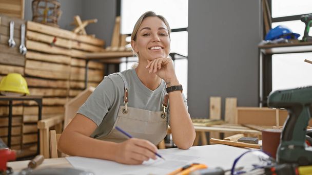 Una mujer sonriente en un taller de carpintería se sienta casualmente con un lápiz, rodeado de herramientas de carpintería y madera. - Foto, Imagen