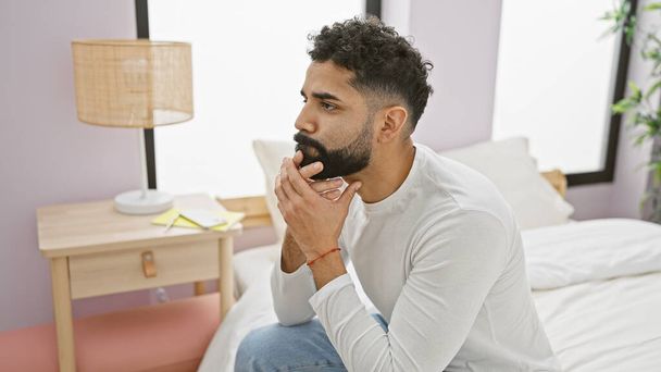 Thoughtful bearded man sitting on the bed in a bedroom looking contemplative, wearing a white shirt and jeans. - Photo, Image
