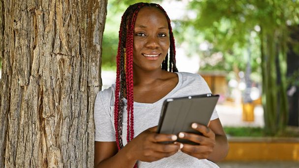 Mulher americana africana confiante, casualmente elegante no parque da cidade, irradia positividade ao usar touchpad, seu sorriso emprestando um ar de alegria infecciosa, cercado pelo verde exuberante ao ar livre. - Foto, Imagem