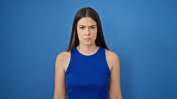 Νεαρή όμορφη ισπανόφωνη γυναίκα στέκεται με σοβαρή έκφραση πάνω από απομονωμένο μπλε φόντο - Φωτογραφία, εικόνα
