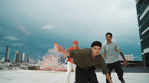 Egy csoport multikulturális hipszter, akik színjelzőt játszanak a tetőn. Vonzó utcai táncos kezében színes füst és végre break tánc lépés hip-hop zene. Kültéri sport 2024. Hatálybalépés. - Fotó, kép