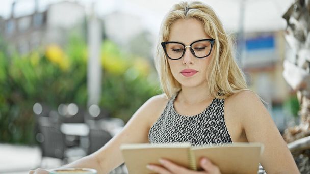 Молодая блондинка сидит на столе с серьезным выражением лица чтение книги на террасе кафе - Фото, изображение