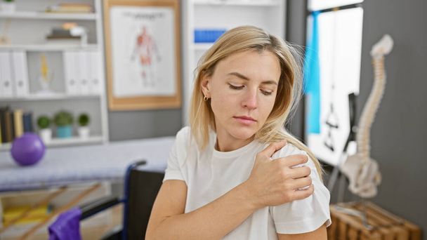 Uma jovem loira mostrando desconforto no ombro em uma clínica de fisioterapia moderna - Foto, Imagem