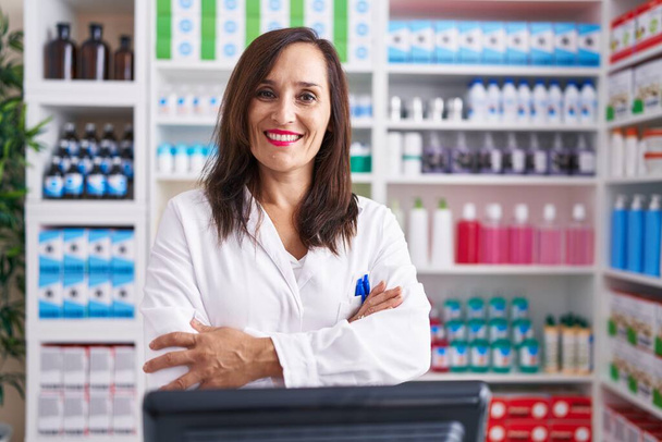 Middelbare leeftijd brunette vrouw werkt bij apotheek drogisterij gelukkig gezicht glimlachend met gekruiste armen kijken naar de camera. positieve persoon.  - Foto, afbeelding