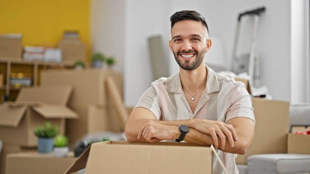 Jovem hispânico sorrindo confiante inclinado em caixa de papelão em nova casa - Foto, Imagem