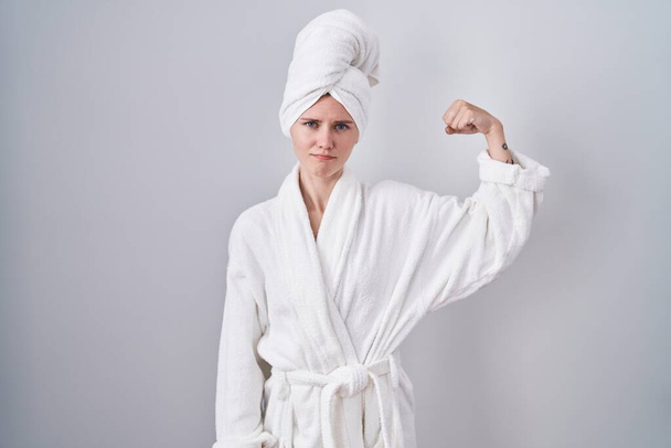 Blonde femme caucasienne portant peignoir personne forte montrant muscle du bras, confiant et fier de la puissance  - Photo, image