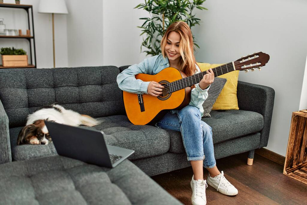 Beyaz bir kadın online klasik gitar dersi alıyor. Evde köpekle birlikte kanepede oturuyor. - Fotoğraf, Görsel