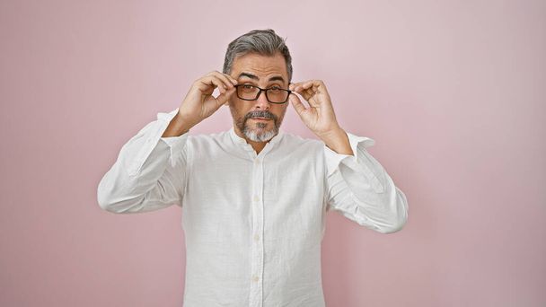 Attraktiver, entspannter junger hispanischer Mann mit grauen Haaren, lässig Brille, modisch vor rosa Hintergrund stehend. Kühle Miene, ernster und doch konzentrierter Blick. - Foto, Bild