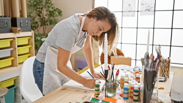 Çekici sarışın kadın sanatçı, odaklanmış yüz çizim defteriyle meşgul, kapalı mekan sanat stüdyosu. - Fotoğraf, Görsel