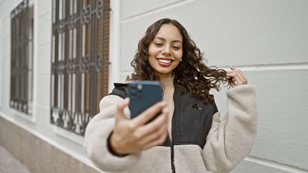 Une jeune femme souriante prend un selfie dans une rue de la ville, mettant en valeur le style de vie urbain moderne et l'utilisation de la technologie. - Photo, image