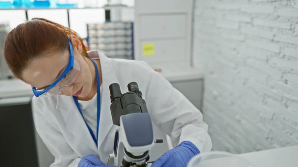 Uma mulher caucasiana focada usando óculos de proteção examina um espécime através de um microscópio em um ambiente de laboratório interno. - Foto, Imagem