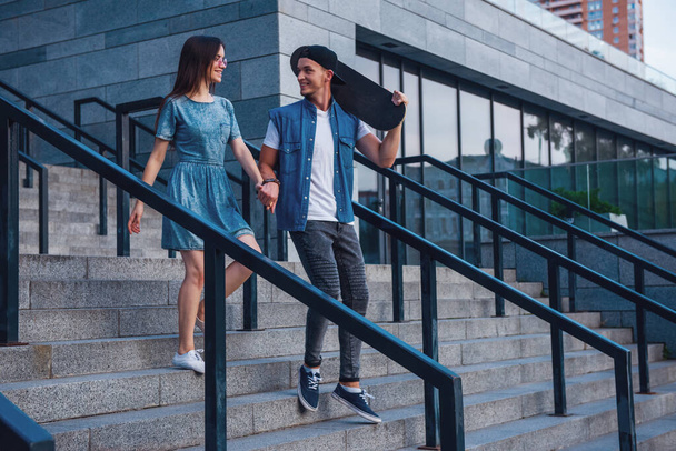 Stylish young couple is holding hands and smiling while walking outdoors, guy is holding a skateboard - Photo, Image