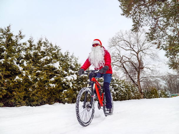Papai Noel monta uma bicicleta no inverno. Um cara com barba e usando um chapéu de Papai Noel cavalga através da neve profunda em uma bicicleta vermelha - Foto, Imagem