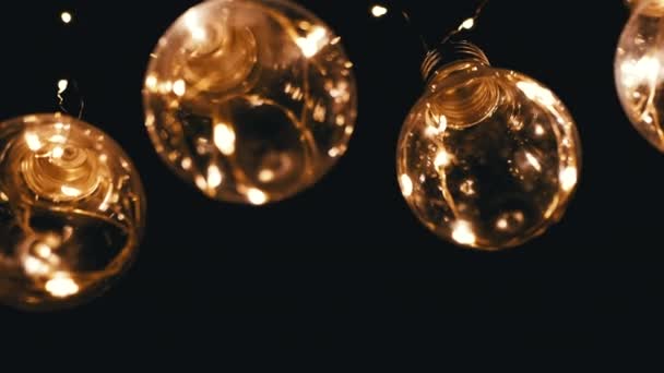 Багато висячих світяться вінтажні лампочки Едісон на чорному тлі. Вид знизу. Тепле світло. Текстура, структура. Рядок. Декоративні старовинні електричні лампочки, гірлянда в стилі ретро освітлюють простір. - Кадри, відео