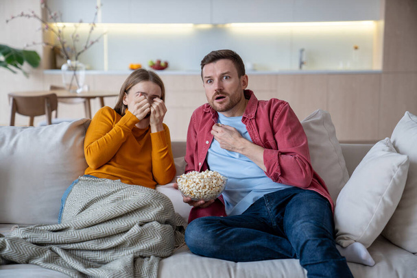Przerażona żona i mąż oglądają thriller jedzący popcorn na kanapie w domu. Obawiam się, że rodzina lubi horrory. Straszna kobieta zamykająca twarz dłońmi podczas oglądania strasznego odcinka filmu - Zdjęcie, obraz