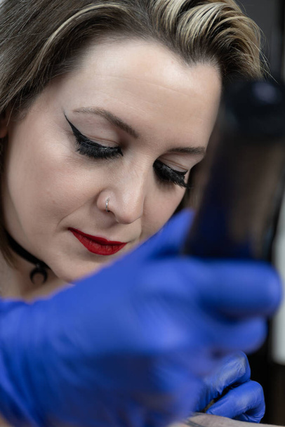 Verticale foto een tatoeëerder met zorgvuldige make-up en rode lippenstift werkt intensief op een tatoeage, presentatie van haar precisie en focus. Concept business, kunst. - Foto, afbeelding