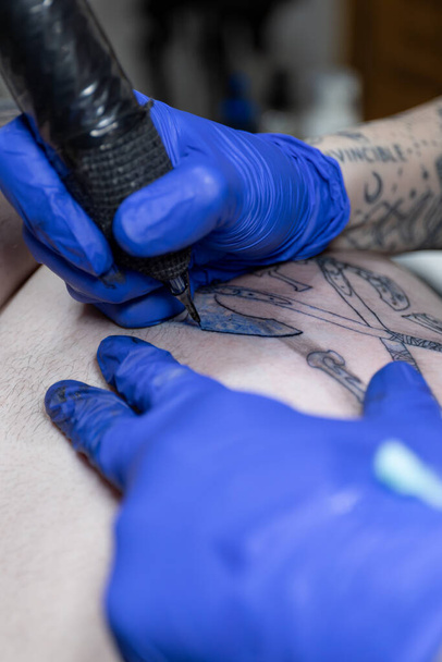 Függőleges fotó A fókuszált lövés megragadja egy tetováló művész kezét, kék kesztyűbe öltözve, hozzáértően részletezve egy tetoválást egy professzionális tetováló géppel. Koncepció üzlet, művészet. - Fotó, kép