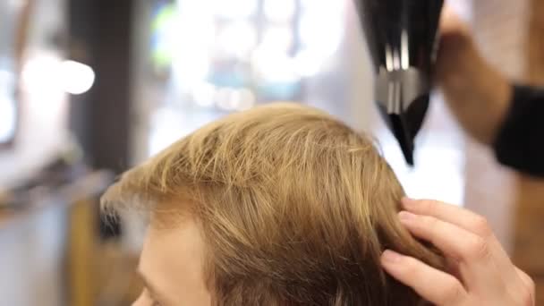 Close up professzionális borbély szárítja man szőke haj hajszárítóval és fésűvel vágás és mosás után a fodrászatban. A fodrász modern férfi frizurát hoz létre a szalonban. Ügyfélszolgálat. Szépségkoncepció - Felvétel, videó
