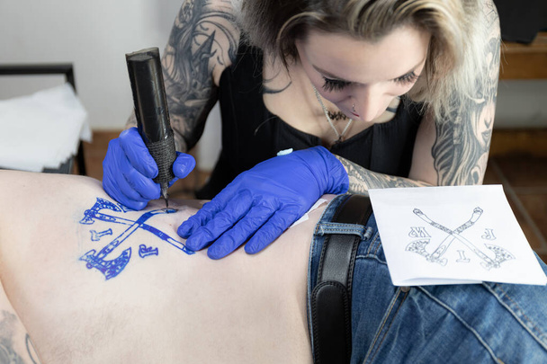 L'artista orizzontale del tatuaggio della foto inchioda meticolosamente uno stemma sul corpo, usando uno schizzo come guida per l'intricato lavoro. Concetto di business, arte. - Foto, immagini
