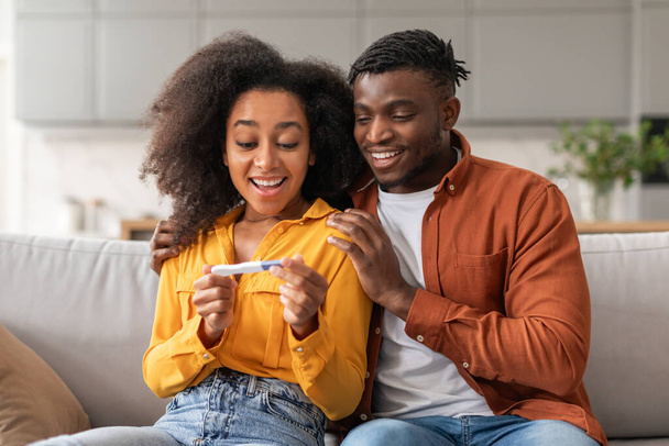 Buď rodiči. Radostné ženatý afroameričan pár držení pozitivní těhotenský test, sedí na pohovce spolu, slaví a objímání v moderním obývacím pokoji doma. Rodinné chvíle, porod - Fotografie, Obrázek