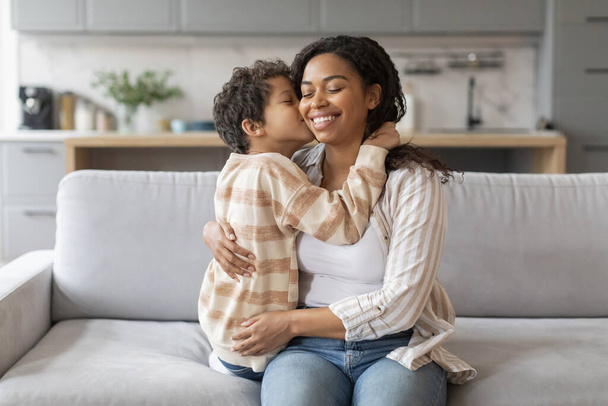 Äitienpäivän konsepti. Musta pieni poika suutelee äitiään poskessa, onnellinen afrikkalainen amerikkalainen äiti ja teini-ikäinen poika sitoutuvat yhteen kotona, nauttivat hellistä hetkistä, rentoutuvat sohvalla olohuoneessa - Valokuva, kuva