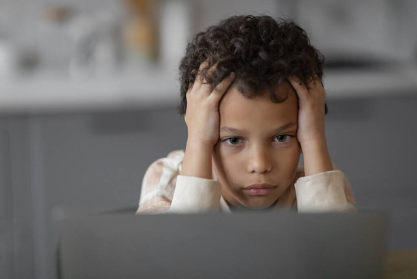 Μικρό μαύρο αγόρι αισθάνεται άγχος και ανησυχία, κρατώντας το κεφάλι του στα χέρια, ενώ κοιτάζοντας οθόνη laptop, αναστατωμένος αρσενικό παιδί κατακλύζεται με εικονική μάθηση ή σε απευθείας σύνδεση δραστηριότητες, closeup - Φωτογραφία, εικόνα