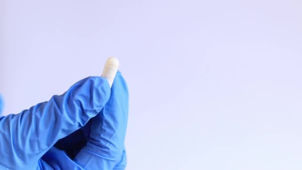 La mano de una mujer en un guante médico azul contiene cápsulas blancas, tabletas. Inspección de una cápsula blanca por un farmacéutico, de cerca. El doctor tiene pastillas. Concepto de medicina y salud - Imágenes, Vídeo