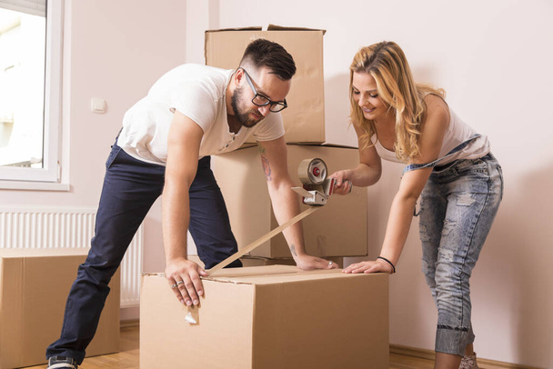 Young girl moving in a new apartment with her boyfriend,standing surrounded with cardboard boxes, packing and taping boxes, while the boyfriend carries boxes away - Photo, Image