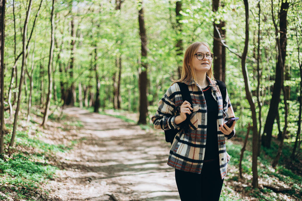 Mladá šťastná žena s batohem pomocí chytrého telefonu a procházky v lese. Jaro. Třicátnice ve sportovním oblečení, turistika po lese. Wanderlust cestovní koncept - Fotografie, Obrázek