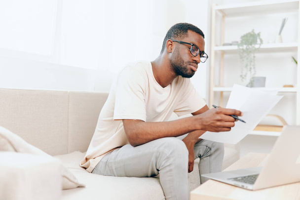 Freelancer afroamericano sonriente que trabaja en el ordenador portátil en la sala de estar moderna o joven afroamericano que escribe en el ordenador portátil, disfrutando de su trabajo independiente mientras está sentado en el sofá en el hogar acogedor - Foto, imagen