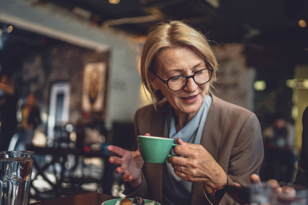 volwassen blanke vrouw zitten in serene sfeer van een gezellige donkere cafe met een kopje koffie in de hand dragen bril genieten van het moment echte persoon kopiëren ruimte - Foto, afbeelding