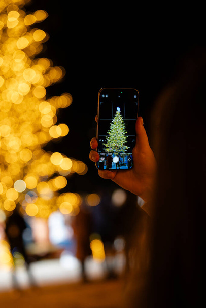 Primer plano de una mano de personas sosteniendo un teléfono inteligente, tomando una foto de un árbol de Navidad bellamente iluminado contra un telón de fondo de luces bokeh doradas.  - Foto, imagen