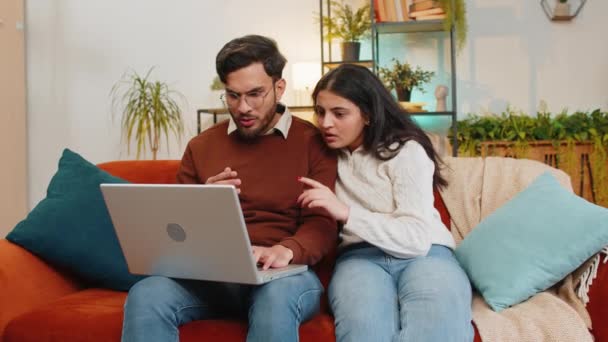 Zszokowana młoda, zróżnicowana para używająca laptopa i przykrywająca usta siedząc na kanapie w domu. zdenerwowany rozczarowany indyjski rodzina mężczyzna kobieta razem czytanie złe wieści nie przegrać na netbook w mieszkaniu. - Materiał filmowy, wideo