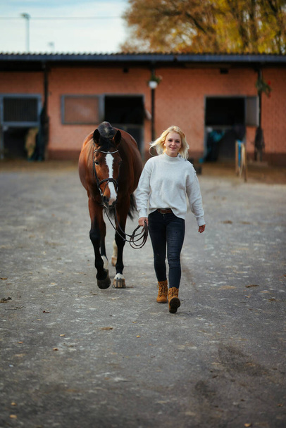 Jeune femme blonde en civil conduit son cheval à travers l'écurie vers la caméra et sourit à son cheval. - Photo, image