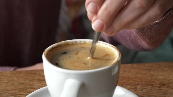 Mão agitando um café Americano em uma pequena xícara de cerâmica branca com uma colher de prata em uma mesa de madeira rústica, tiro de close-up. - Filmagem, Vídeo