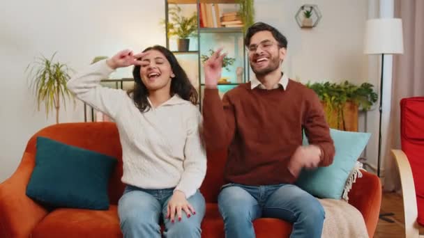Fröhlich aufgeregte junge indische Tanzpaare geben High Five, die auf dem Sofa im heimischen Wohnzimmer sitzen. Lächelnd glücklich diverse Freundin und Freund genießen Wochenende zusammen auf Couch in Wohnung. - Filmmaterial, Video