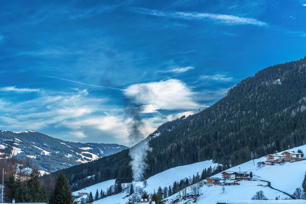 Un paesaggio invernale sereno che mostra un villaggio di montagna innevato, con fumo che sorge sullo sfondo di un cielo azzurro. Ideale per concetti di tranquillità, mete turistiche e bellezze naturali - Foto, immagini