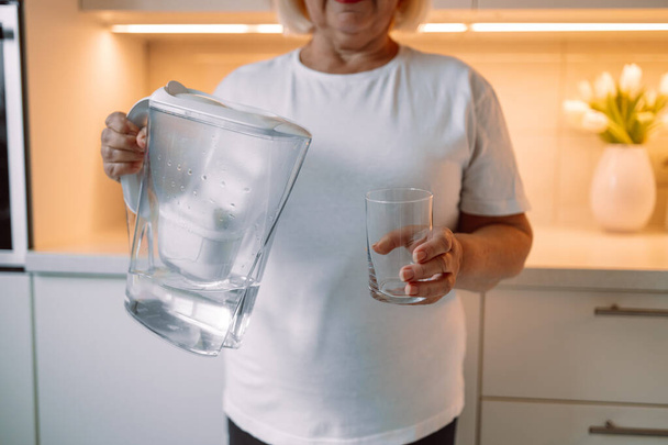 Sorridente donna anziana versando acqua filtrata nel vetro, interno della cucina. Attraente donna anziana in abito casual in possesso di vetro di acqua dolce, spazio copia. Foto di alta qualità - Foto, immagini